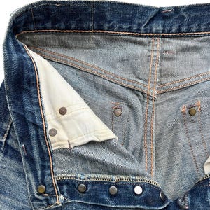 Pu&ograve; includere: Primo piano della parte superiore di jeans blu in denim, che mostra la chiusura con bottoni, i rivetti metallici e i dettagli delle cuciture. I jeans hanno un aspetto usurato e sbiadito, con segni di usura visibili.