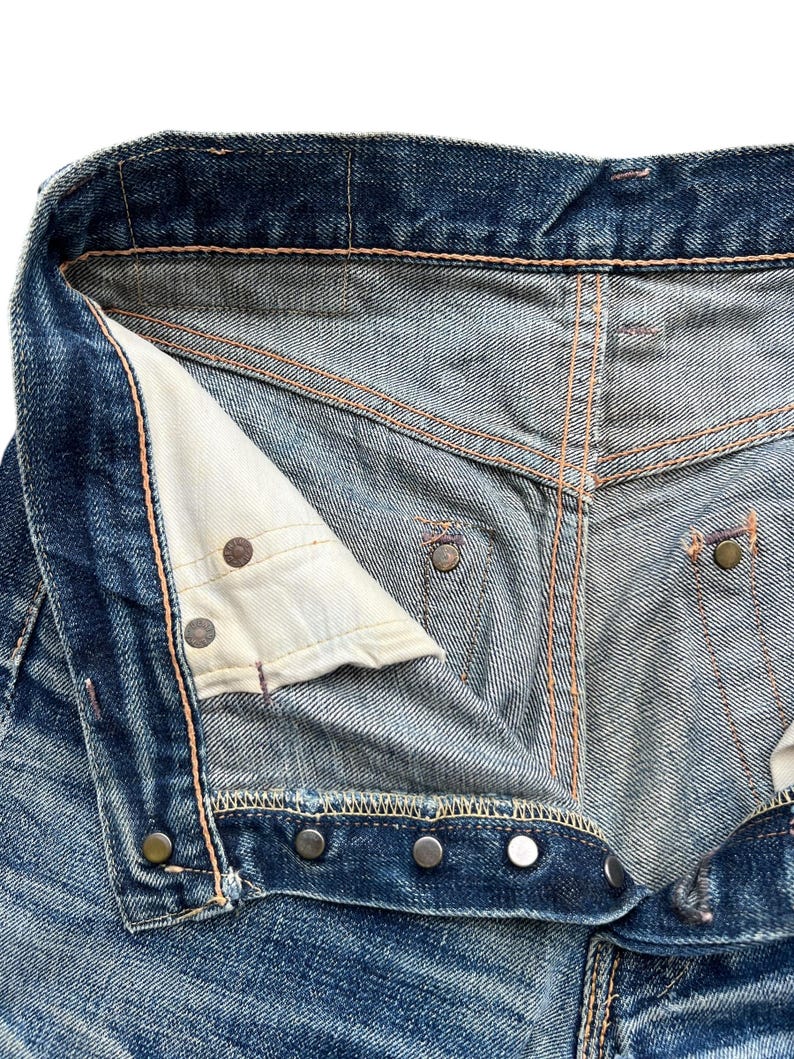 Pu&ograve; includere: Primo piano della parte superiore di jeans blu in denim, che mostra la chiusura con bottoni, i rivetti metallici e i dettagli delle cuciture. I jeans hanno un aspetto usurato e sbiadito, con segni di usura visibili.