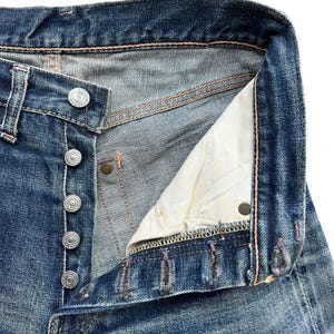 Pu&ograve; includere: Primo piano di jeans di denim blu con abbottonatura. I jeans mostrano segni di usura e sbiadimento, con cuciture e bottoni metallici visibili. La fodera interna &egrave; di colore beige chiaro.