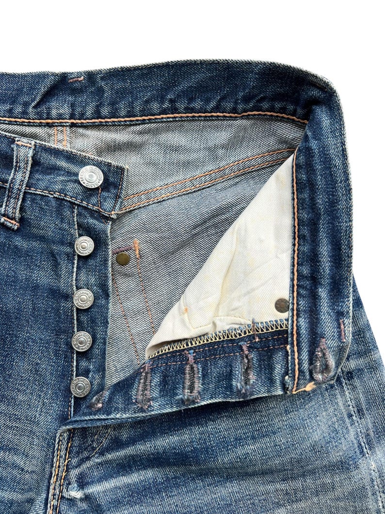 Pu&ograve; includere: Primo piano di jeans di denim blu con abbottonatura. I jeans mostrano segni di usura e sbiadimento, con cuciture e bottoni metallici visibili. La fodera interna &egrave; di colore beige chiaro.