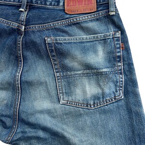Puede incluir: Primer plano de la parte trasera de unos vaqueros azules. Los vaqueros tienen un lavado descolorido y un bolsillo trasero rectangular. Un parche de cuero marr&oacute;n con la marca "EDWIN" es visible en la cintura. Los vaqueros est&aacute;n etiquetados "505X L 34 34".