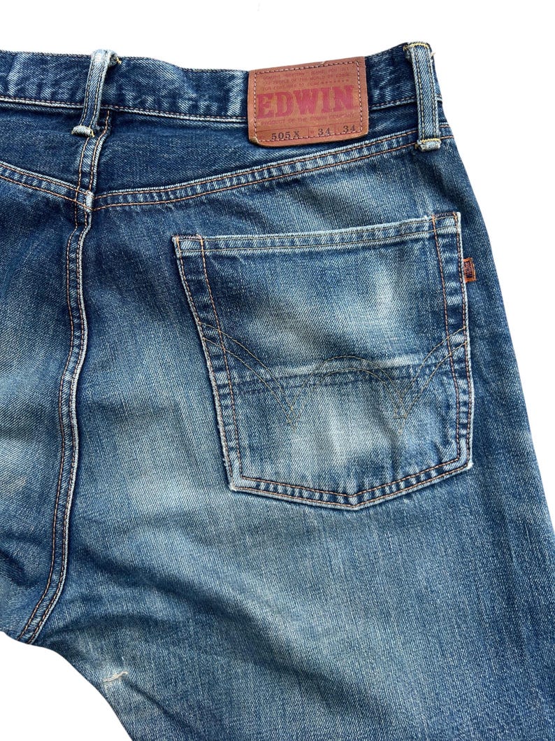 Puede incluir: Primer plano de la parte trasera de unos vaqueros azules. Los vaqueros tienen un lavado descolorido y un bolsillo trasero rectangular. Un parche de cuero marr&oacute;n con la marca "EDWIN" es visible en la cintura. Los vaqueros est&aacute;n etiquetados "505X L 34 34".