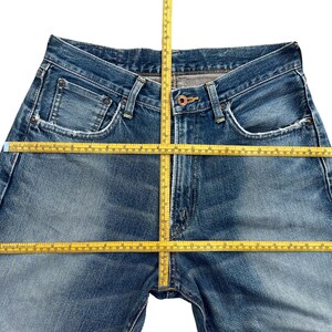 Puede incluir: Primer plano de unos vaqueros azules con cierre de bot&oacute;n y bolsillos. Los vaqueros est&aacute;n planos, con una cinta m&eacute;trica amarilla en la cintura y las piernas. El denim tiene un lavado descolorido.