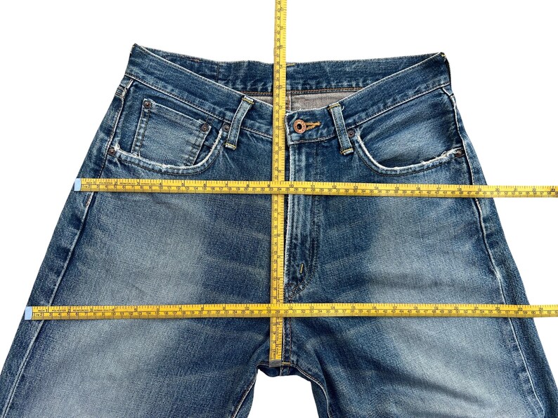 Puede incluir: Primer plano de unos vaqueros azules con cierre de bot&oacute;n y bolsillos. Los vaqueros est&aacute;n planos, con una cinta m&eacute;trica amarilla en la cintura y las piernas. El denim tiene un lavado descolorido.