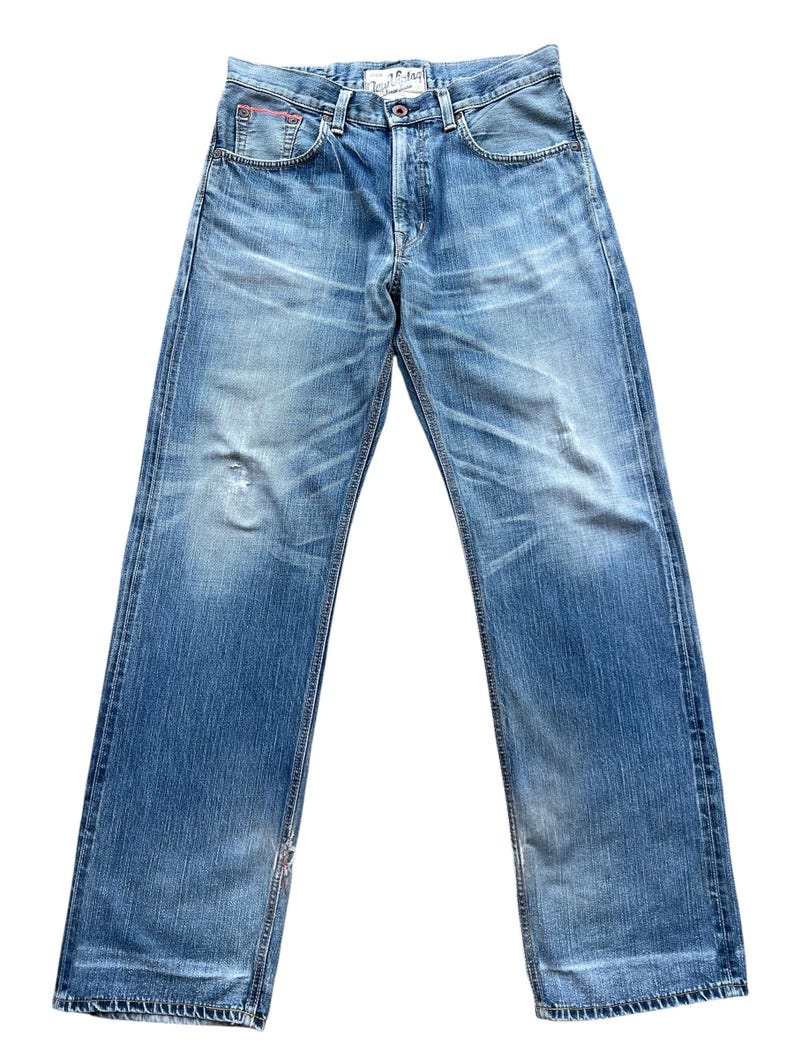 Pu&ograve; includere: Jeans di denim blu sbiadito con un classico design a cinque tasche. I jeans hanno una chiusura con bottoni e un aspetto invecchiato con sbiadimento e segni di usura. La vita presenta un piccolo accento rosso. I jeans sono un modello a gamba dritta.