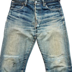 May include: Faded blue denim jeans with a button fly and classic five-pocket design. The jeans show signs of wear with distressed areas and patches, giving them a vintage look. The denim has a worn-in appearance.