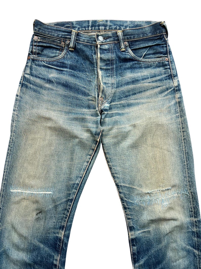 May include: Faded blue denim jeans with a button fly and classic five-pocket design. The jeans show signs of wear with distressed areas and patches, giving them a vintage look. The denim has a worn-in appearance.