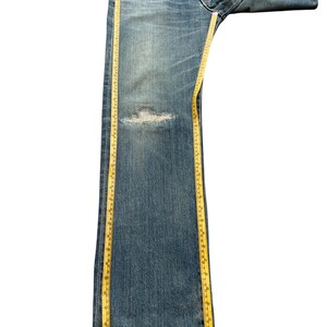 May include: Blue denim jeans with a distressed knee area. A yellow measuring tape is placed along the length of the jeans. The jeans are folded over at the waist.