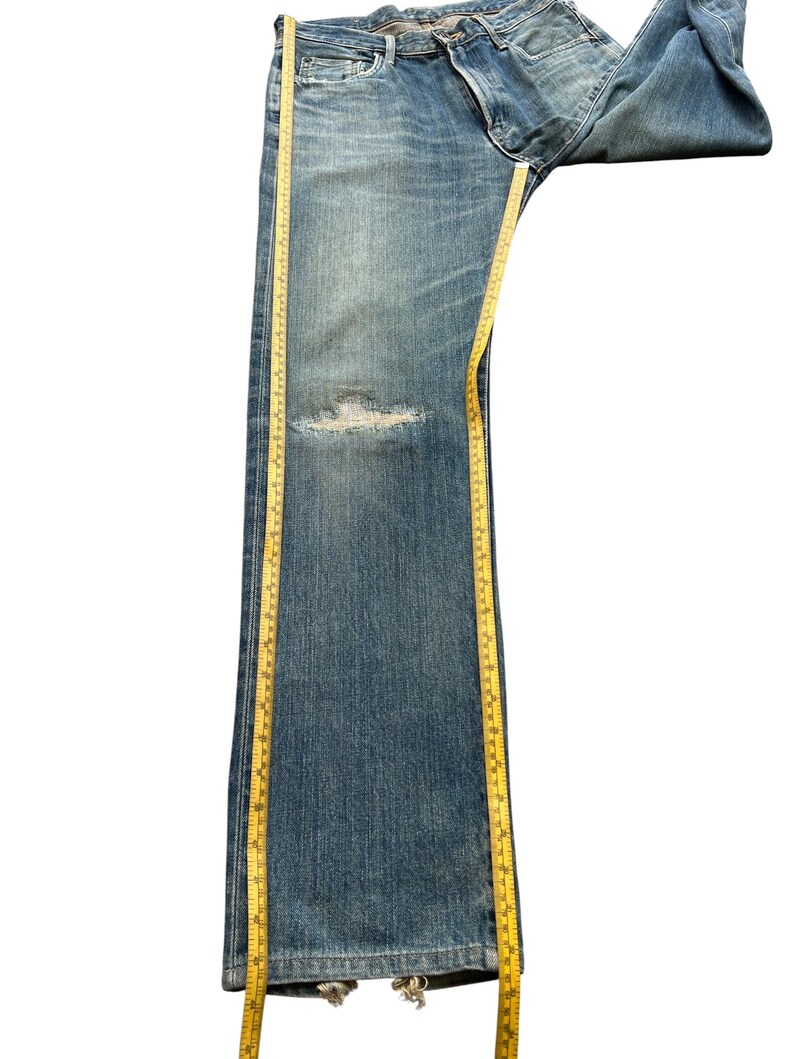 May include: Blue denim jeans with a distressed knee area. A yellow measuring tape is placed along the length of the jeans. The jeans are folded over at the waist.
