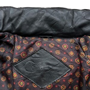 May include: Close-up of a black leather jacket interior. The lining is a dark brown fabric with a repeating pattern of red and gold medallions. A black leather patch with a decorative edge is sewn into the lining.