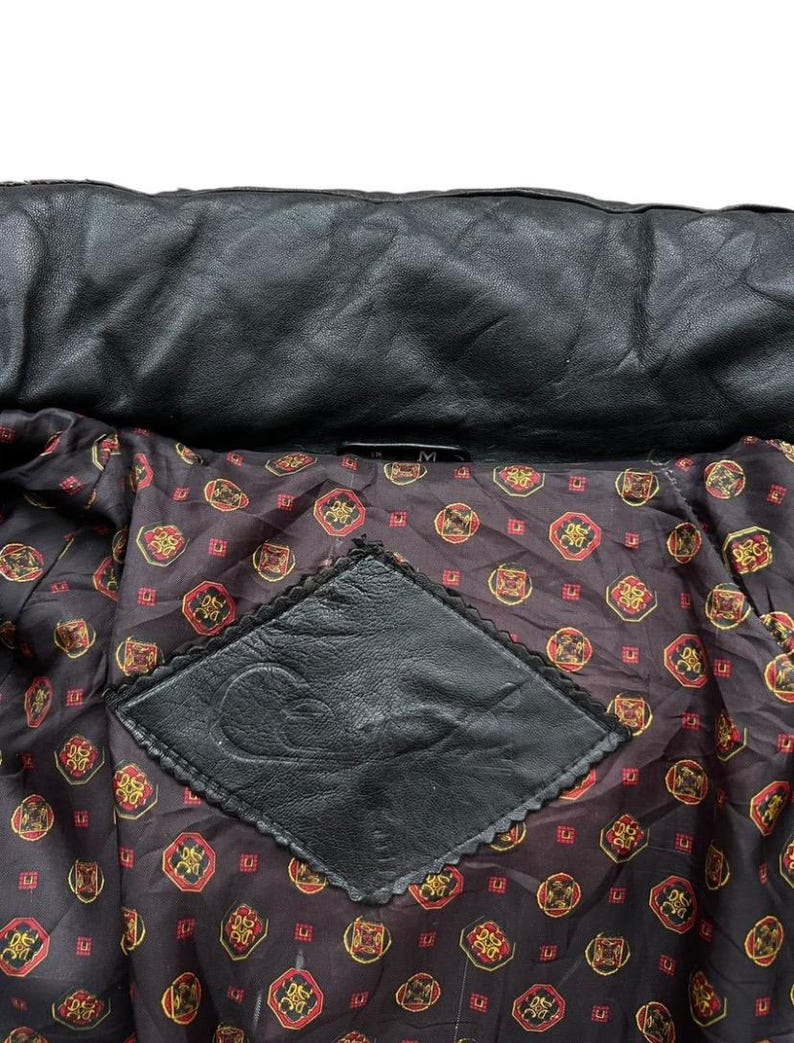 May include: Close-up of a black leather jacket interior. The lining is a dark brown fabric with a repeating pattern of red and gold medallions. A black leather patch with a decorative edge is sewn into the lining.