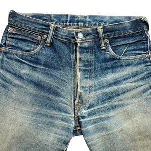 May include: Close-up of blue denim jeans with a button fly. The jeans show signs of wear and fading, with visible pocket details and rivets. The denim has a classic, vintage look.