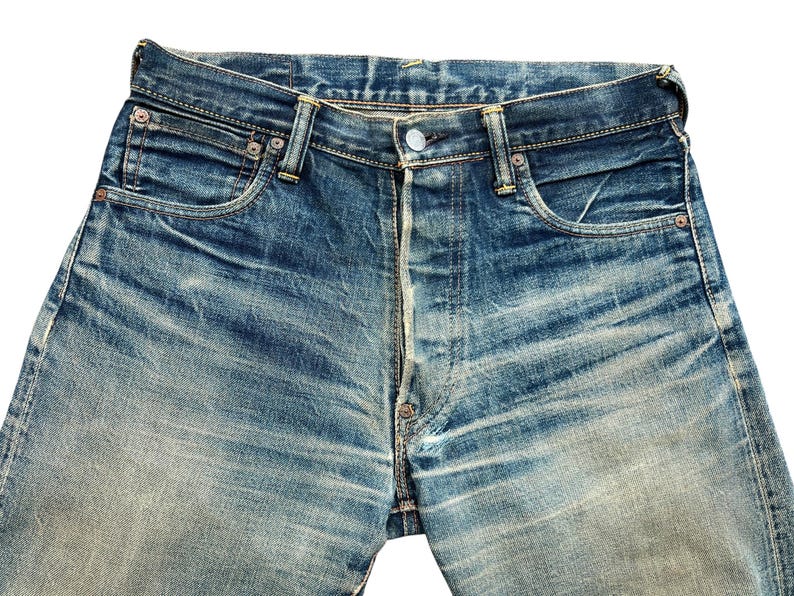 May include: Close-up of blue denim jeans with a button fly. The jeans show signs of wear and fading, with visible pocket details and rivets. The denim has a classic, vintage look.