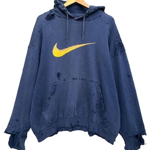Puede incluir: Una sudadera azul desgastada con un logotipo de Nike swoosh amarillo. La sudadera tiene varios agujeros y desgarros.