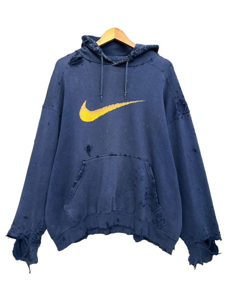 Puede incluir: Una sudadera azul desgastada con un logotipo de Nike swoosh amarillo. La sudadera tiene varios agujeros y desgarros.