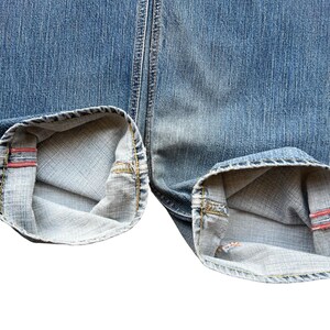 Pu&ograve; includere: Primo piano dei risvolti ripiegati di jeans di denim blu. I risvolti sono ripiegati per rivelare il tessuto interno e un dettaglio di bordo rosso. I jeans hanno un aspetto classico e usurato.