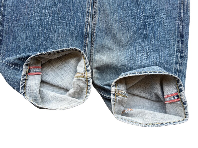Pu&ograve; includere: Primo piano dei risvolti ripiegati di jeans di denim blu. I risvolti sono ripiegati per rivelare il tessuto interno e un dettaglio di bordo rosso. I jeans hanno un aspetto classico e usurato.