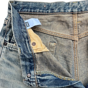 May include: Close-up of blue denim jeans with a partially unbuttoned fly. The jeans show signs of wear and fading, with visible stitching and a small tag. The zipper is partially open, revealing the interior lining.