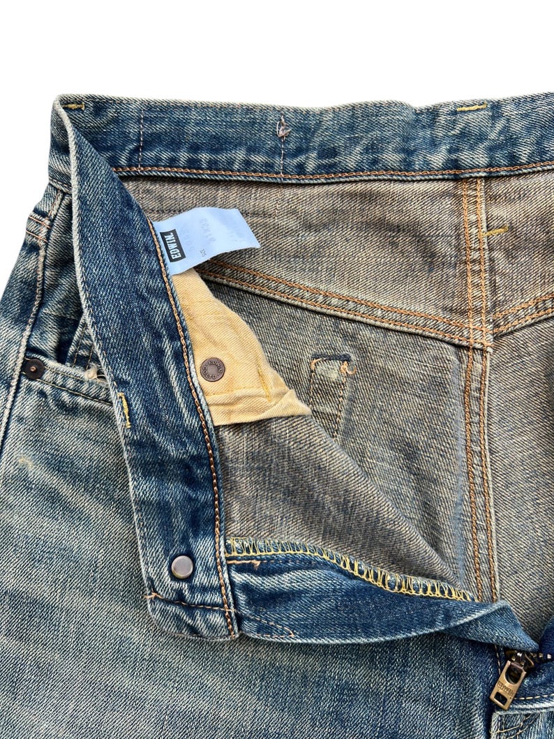 May include: Close-up of blue denim jeans with a partially unbuttoned fly. The jeans show signs of wear and fading, with visible stitching and a small tag. The zipper is partially open, revealing the interior lining.