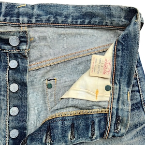 May include: Close-up of vintage blue denim jeans, showing the button fly, metal buttons, and stitching details. The jeans have a worn, faded appearance, with a small tag visible inside. The fabric is 100% cotton.