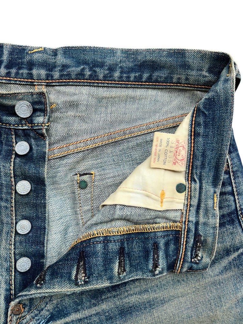 May include: Close-up of vintage blue denim jeans, showing the button fly, metal buttons, and stitching details. The jeans have a worn, faded appearance, with a small tag visible inside. The fabric is 100% cotton.