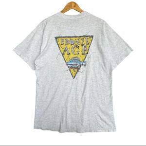 Op de afbeelding: Een lichtgrijs t-shirt met korte mouwen en een afbeelding op de achterkant. De afbeelding toont een gele driehoek met gebarsten randen met de tekst BRONZE AGE in zwarte letters boven een blauwe visgraatillustratie op een witte achtergrond.