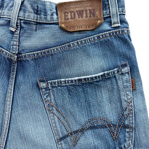 Pu&ograve; includere: Primo piano di jeans blu in denim con un'etichetta in pelle marrone con il marchio "EDWIN" e testo. I jeans hanno una tasca posteriore con cuciture decorative e una piccola etichetta marrone. Il testo "MADE IN JAPAN" &egrave; visibile.