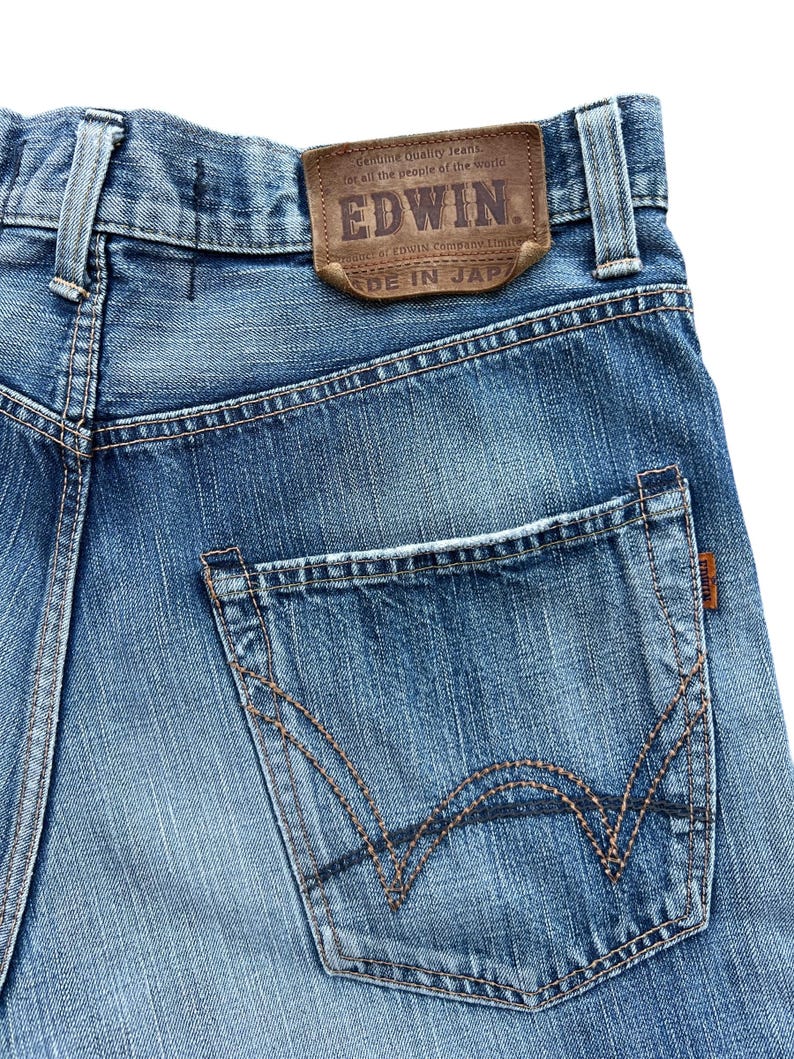 Pu&ograve; includere: Primo piano di jeans blu in denim con un'etichetta in pelle marrone con il marchio "EDWIN" e testo. I jeans hanno una tasca posteriore con cuciture decorative e una piccola etichetta marrone. Il testo "MADE IN JAPAN" &egrave; visibile.