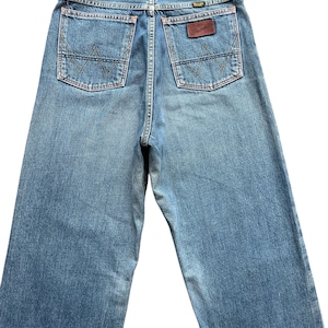 May include: Light blue denim jeans with the Wrangler logo on the back pocket. The jeans have two back pockets with a stitched design and a brown leather patch with the brand name.