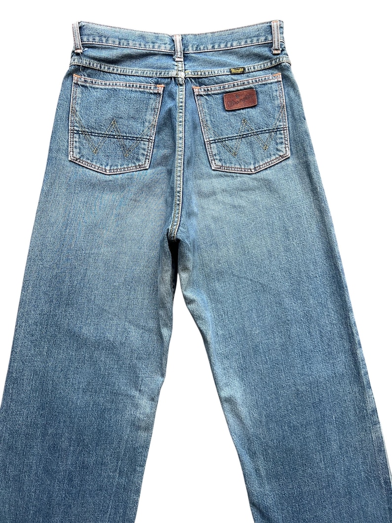 May include: Light blue denim jeans with the Wrangler logo on the back pocket. The jeans have two back pockets with a stitched design and a brown leather patch with the brand name.