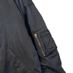 May include: Close-up of a black bomber jacket. The jacket features a zippered sleeve pocket with a gold-colored zipper and a snap-closure pocket. The fabric appears to be a smooth, matte material.