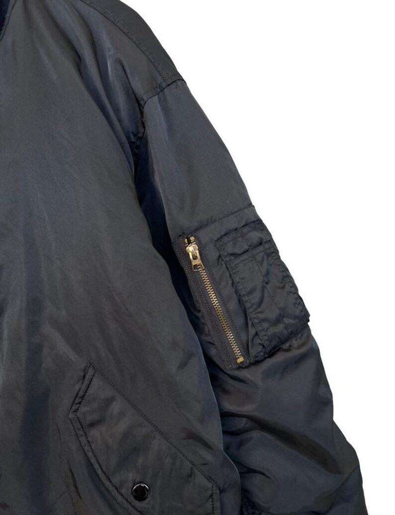 May include: Close-up of a black bomber jacket. The jacket features a zippered sleeve pocket with a gold-colored zipper and a snap-closure pocket. The fabric appears to be a smooth, matte material.