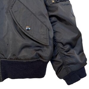 May include: Close-up of a black bomber jacket. The jacket features a dark navy blue ribbed hem and cuffs. A pocket with a snap closure is visible on the left side. The sleeve is gathered at the elbow.