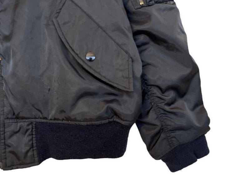 May include: Close-up of a black bomber jacket. The jacket features a dark navy blue ribbed hem and cuffs. A pocket with a snap closure is visible on the left side. The sleeve is gathered at the elbow.