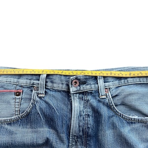 Pu&ograve; includere: Primo piano di jeans blu con un metro a nastro giallo sulla vita. I jeans hanno un classico design a cinque tasche, con un lavaggio sbiadito e dettagli usurati. Il metro a nastro mostra la misura della vita in centimetri.