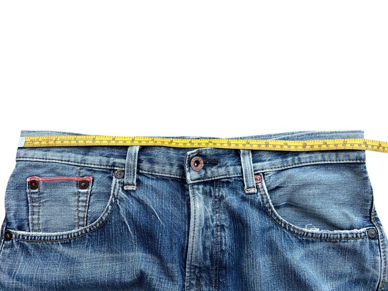 Pu&ograve; includere: Primo piano di jeans blu con un metro a nastro giallo sulla vita. I jeans hanno un classico design a cinque tasche, con un lavaggio sbiadito e dettagli usurati. Il metro a nastro mostra la misura della vita in centimetri.