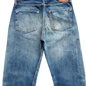 以下が含まれることがあります： 色あせた青色のデニムジーンズの後ろ姿。2つのバックポケットが付いています。ジーンズには革のパッチと、片方のポケットに赤いタブが付いています。デニムは使用感があり、色あせが見られます。