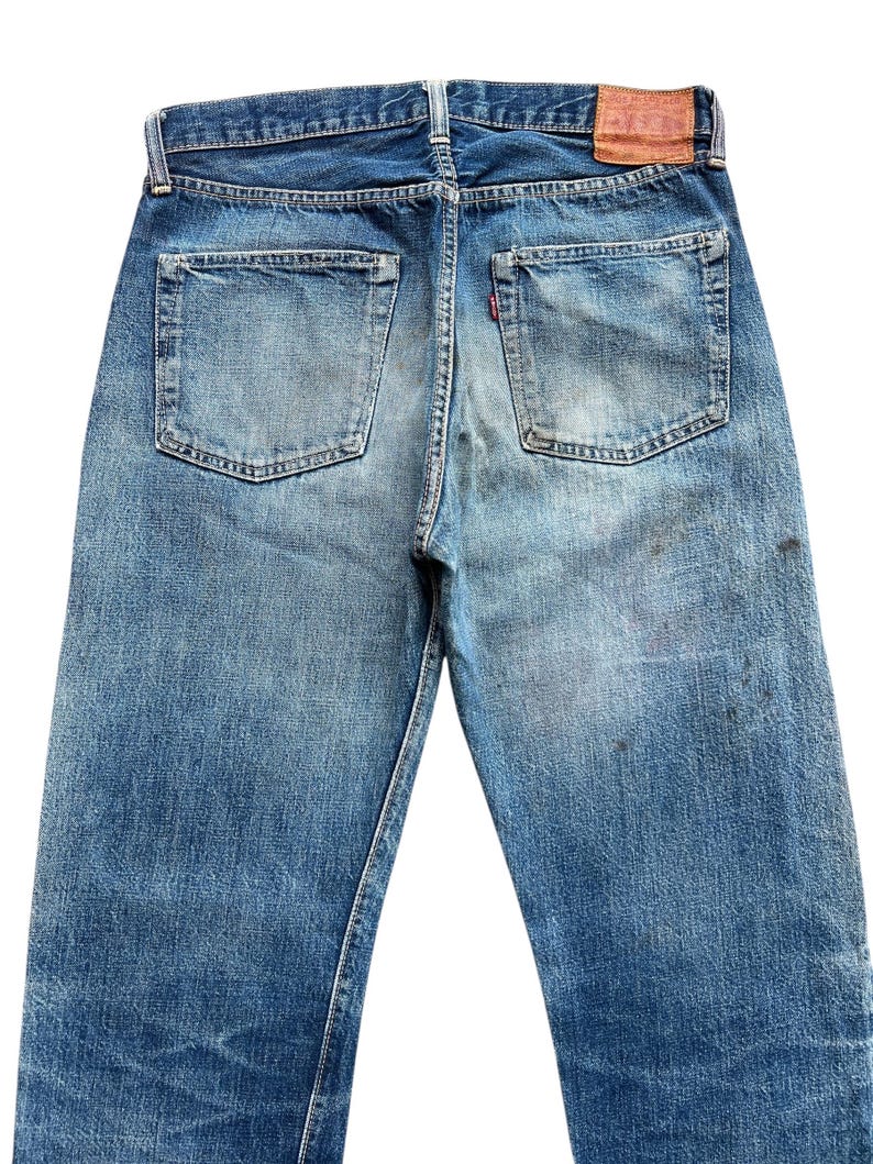 以下が含まれることがあります： 色あせた青色のデニムジーンズの後ろ姿。2つのバックポケットが付いています。ジーンズには革のパッチと、片方のポケットに赤いタブが付いています。デニムは使用感があり、色あせが見られます。
