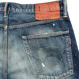 Puede incluir: Primer plano de unos vaqueros de mezclilla azules con aspecto desgastado. El bolsillo trasero es visible, junto con un parche de cuero marr&oacute;n que dice "MADE IN JAPAN". Los vaqueros tienen un dise&ntilde;o cl&aacute;sico de cinco bolsillos.
