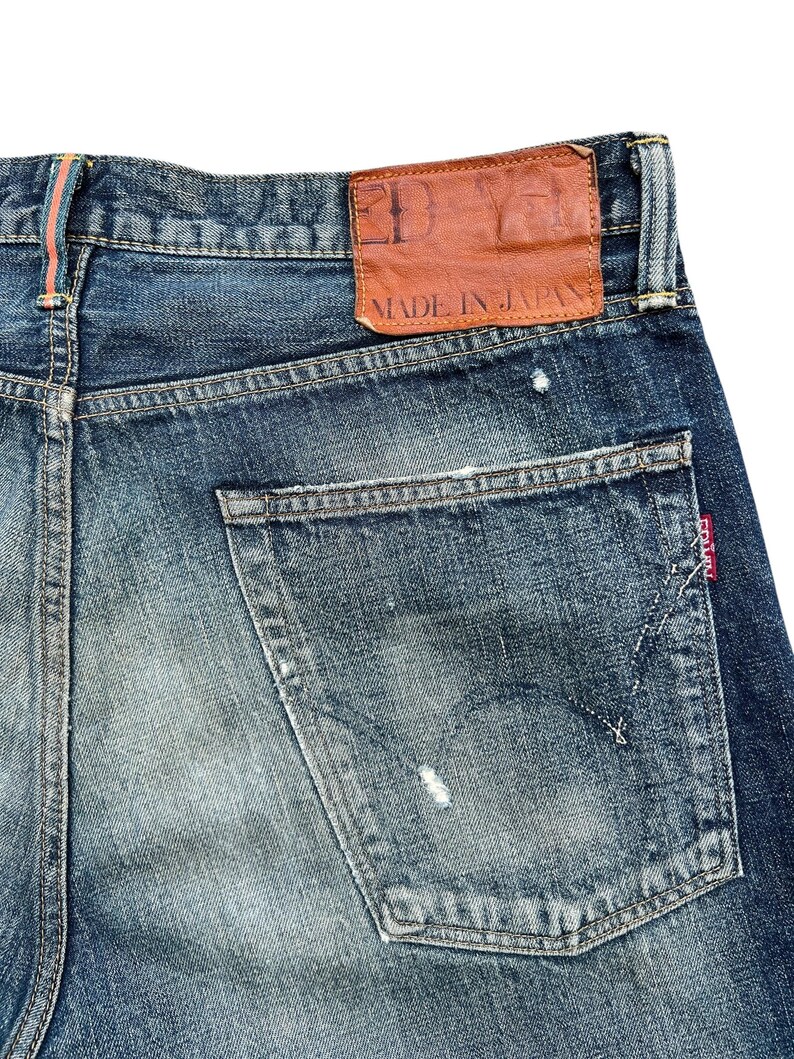 Puede incluir: Primer plano de unos vaqueros de mezclilla azules con aspecto desgastado. El bolsillo trasero es visible, junto con un parche de cuero marr&oacute;n que dice "MADE IN JAPAN". Los vaqueros tienen un dise&ntilde;o cl&aacute;sico de cinco bolsillos.
