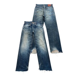 Puede incluir: Dos pares de vaqueros de mezclilla azul vintage. Un par está plano, mostrando el frente, con un aspecto desgastado y una sección rasgada en la rodilla. El otro par está detrás, mostrando la parte trasera. Ambos pares tienen un diseño clásico de cinco bolsillos.