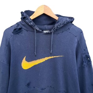 Puede incluir: Una sudadera azul desgastada con un logotipo de Nike swoosh amarillo. La sudadera tiene varios agujeros y desgarros.