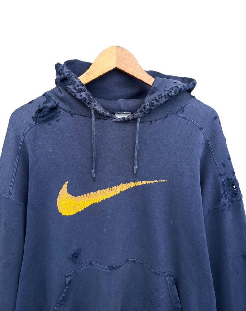 Puede incluir: Una sudadera azul desgastada con un logotipo de Nike swoosh amarillo. La sudadera tiene varios agujeros y desgarros.
