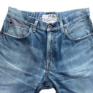 Pu&ograve; includere: Primo piano di jeans di denim blu con un lavaggio sbiadito. I jeans hanno un classico stile a cinque tasche, una chiusura con bottoni e una piccola tasca con un accento rosso. L'etichetta recita "New Vintage Stone Jeans".