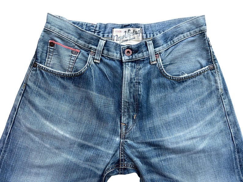 Pu&ograve; includere: Primo piano di jeans di denim blu con un lavaggio sbiadito. I jeans hanno un classico stile a cinque tasche, una chiusura con bottoni e una piccola tasca con un accento rosso. L'etichetta recita "New Vintage Stone Jeans".