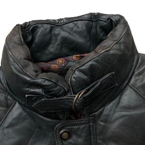 May include: Close-up of a black leather jacket collar. The collar is padded and has a buckle closure. The jacket has a quilted design and a button closure. The interior lining is patterned with red and gold accents.