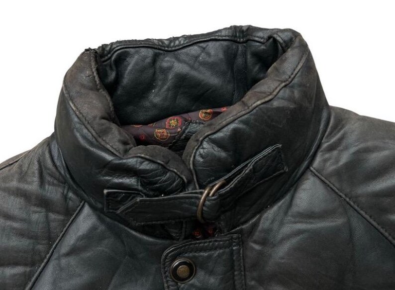 May include: Close-up of a black leather jacket collar. The collar is padded and has a buckle closure. The jacket has a quilted design and a button closure. The interior lining is patterned with red and gold accents.