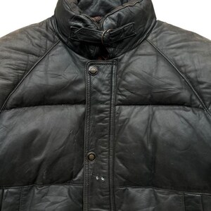 May include: Close-up of a black leather puffer jacket. The jacket features a high collar with a buckle closure, a button-down front, and quilted panels. The leather appears worn, adding character to the garment.