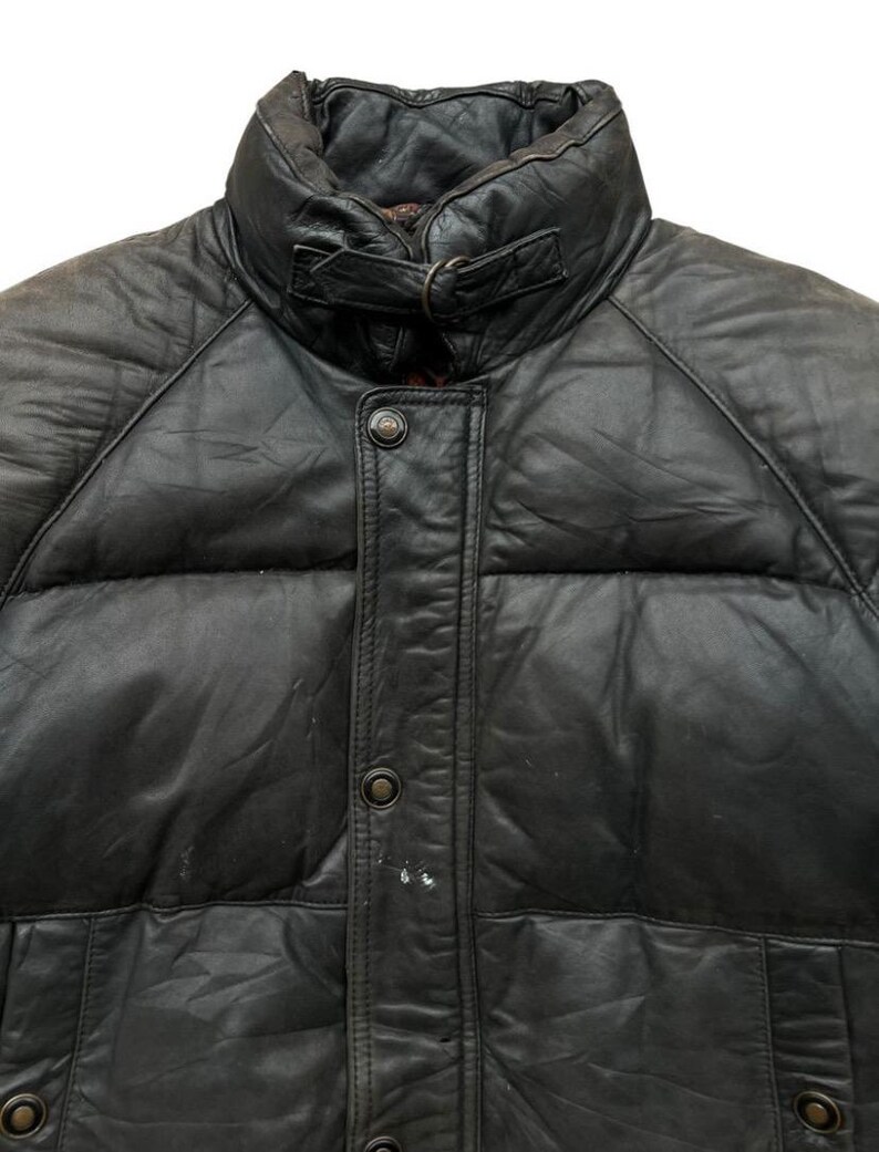 May include: Close-up of a black leather puffer jacket. The jacket features a high collar with a buckle closure, a button-down front, and quilted panels. The leather appears worn, adding character to the garment.