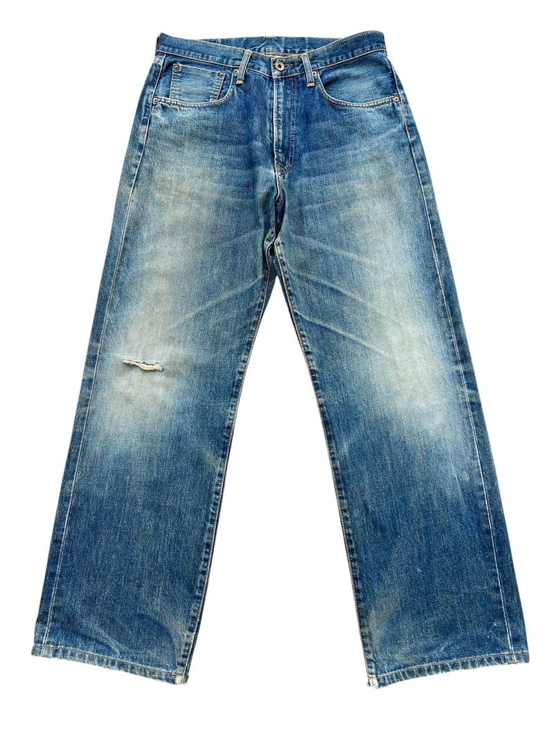 May include: Faded blue denim jeans with a distressed look. The jeans have a classic five-pocket design, a button and zipper closure, and a small tear on the left leg. The jeans have a straight leg cut.